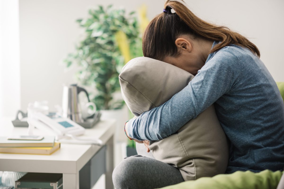 Unhappy lonely depressed woman at home, she is sitting on the couch and hiding her face on a pillow, depression concept